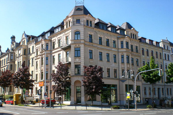 Historisches Mehrfamilienhaus mit Geschäften im Erdgeschoss an einer Straßenecke in einer europäischen Stadt bei sonnigem Wetter.