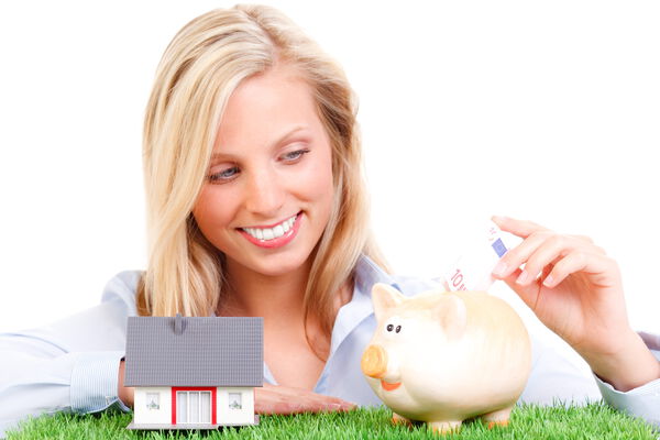 Junge Frau steckt Geldschein in ein Sparschwein, neben einem Miniaturhaus auf einer grünen Wiese – Symbol für Sparen und Eigenheimfinanzierung.